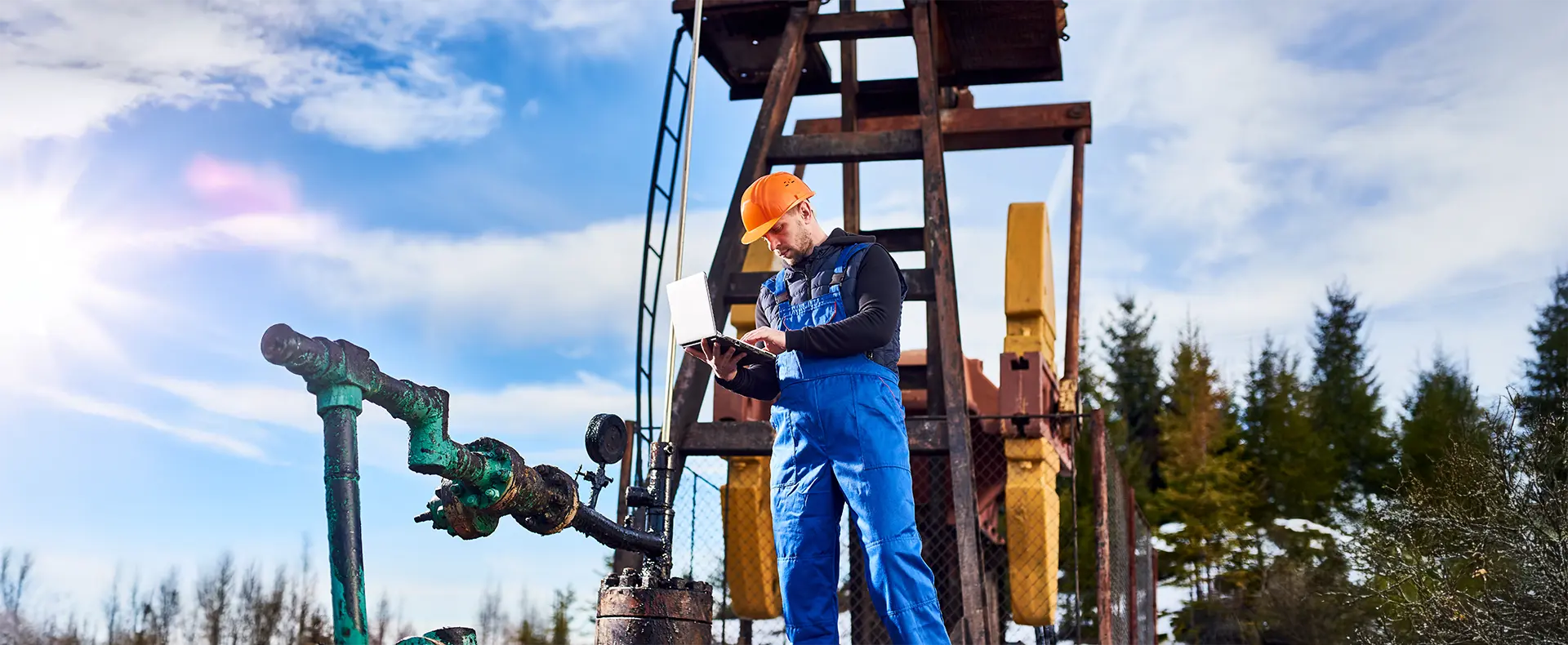 Oil field worker A field worker with an orange safety helmet standing in front of an oil rig and looking down at his laptop