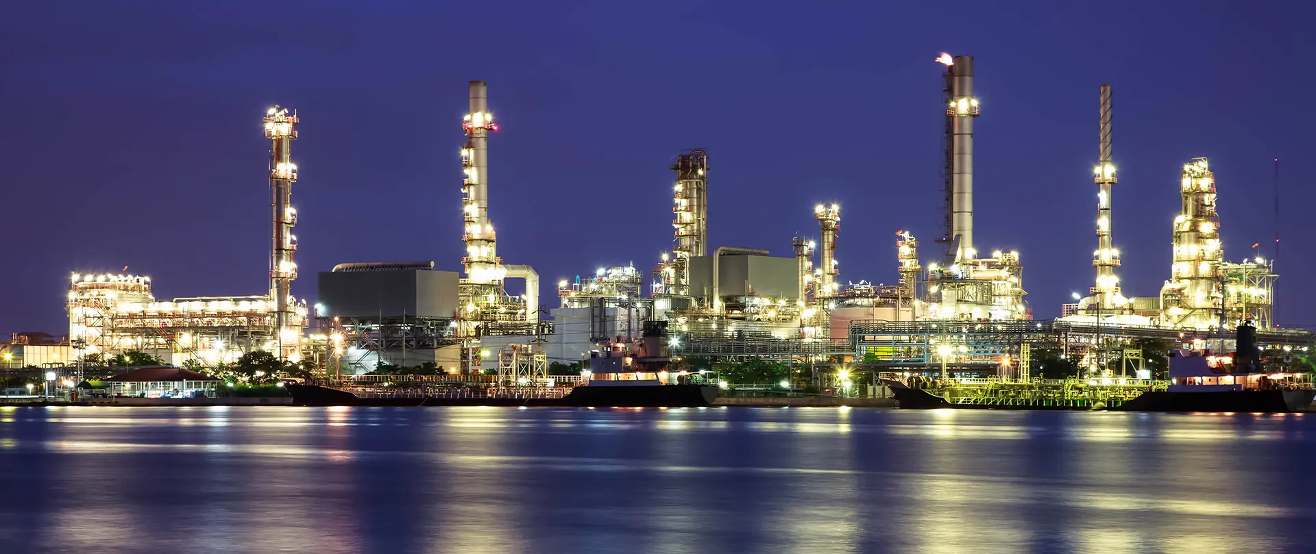 Brightly lit refinery or petrochemical near a waterside at night with chimneys and cargo ships