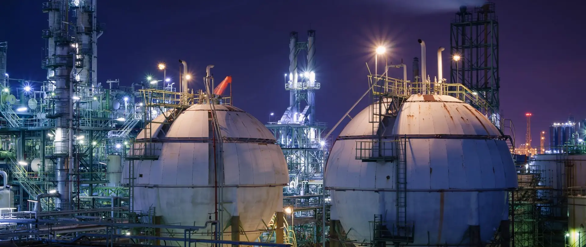 Petrochemical refinery at night with illuminated spherical storage tanks and a complex network of industrial pipes and metal structures