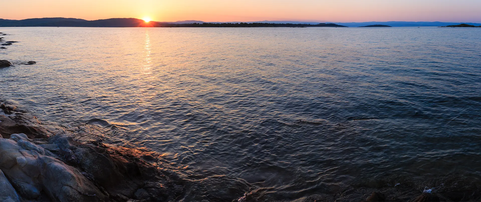 Sunset over calm sea with rocky shoreline and distant hills